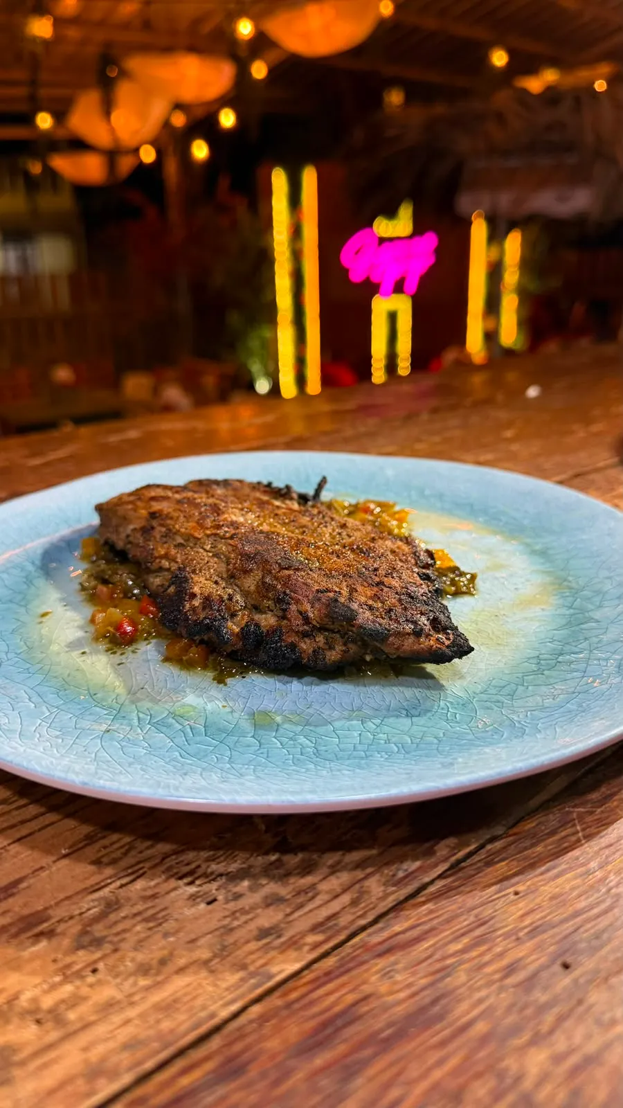 Churrasco a la parrilla sobre vegetales, frente al letrero neón de Sazonao Pop.
